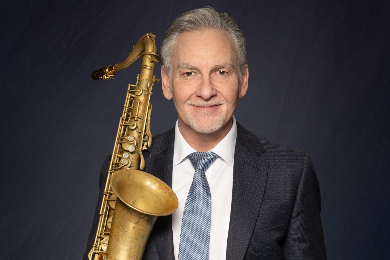 Mann in Anzug hält ein Saxophon, lächelt freundlich in die Kamera.